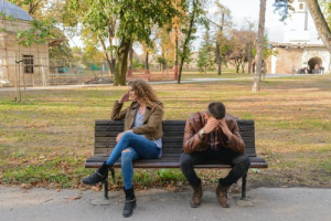 Couple sitting next to each other on a bench in a park. Couple looks like they are mad at each other.