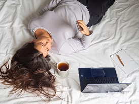 Woman laying in bed looking at cellphone procrastinating work. Lazy girl working and studying from home in bed in the morning with cup of coffee . Relaxing morning , social media addiction. Copy space