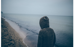 a person standing on a beach in a black hoodie covering his face looking out at the ocean