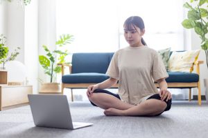 A woman is sitting on the floor in a living room, engaging in yoga to rejuvenate herself while following instructions from her laptop.