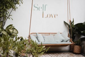 There is a hanging chair in a room adorned with plants, and the phrase self-care is written on the wall, creating a trangquil and inviting space for relaxation well-being