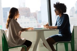 Two women are sitting across from each other, talking to one another. Women may be discussing their problems and/or needs. They may be one another's support system. 