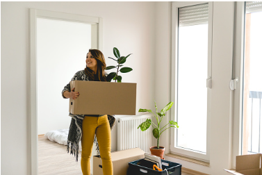Woman is holding a box with a plant sticking out of it. The woman looks like she is moving into  a new apartment. Woman may be experiencing a quarter life crisis. 