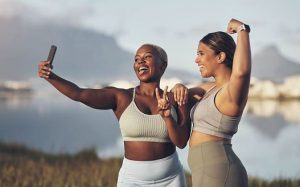 Two women are posing, taking a selfie with each other while wearing workout attire. Both women are practicing techniques that can help you in loving yourself.