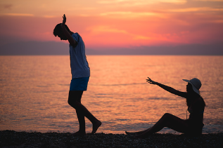 In this image we see a couple sitting at a beach scenery. The woman is sitting on the floor with her arm reaching towards the man as he is walking away from her. Couples therapy is available virtually or in person at our Woodland Hills, CA therapy practice.