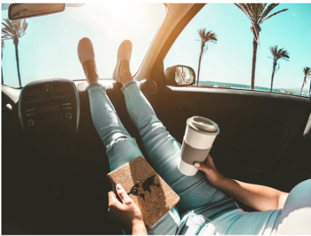 In this image we see a woman sitting in the passenger seat of her car, with her legs up on the dashboard, holding a notebook and a cup of coffee. Outside we see palm trees under the blue sky. Individual therapy is available at our Woodland Hills, CA therapy practice for virtual or in person sessions 91364 | 91307 | 91356