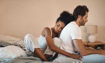 This image shows a couple sitting on the bed together. The woman in the image is sitting in bed hugging the man from behind with her arms around him. The man is sitting on the edge of the bed with his feet on the floor. Both of their faces are sad which shows they are struggling with their mental health. Couples therapy is available at our Woodland Hills therapy office. 91364 | 91307 | 91356