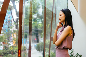 A woman is standing in front of glass windows, looking outside, with her hand on her chin. She looks like she is in deep thought about something, possibly experiencing shame or guilt about something in her life. CBT helps give you your power back and work towards being stress free, less fatigued, and happier. CBT therapy offered at Woodland Hills therapy office. 