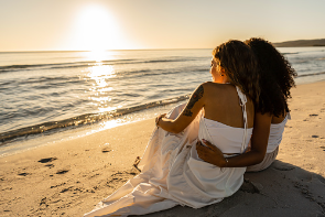Two women are sitting on the beach wearing white dresses, hugging one another. Couples therapy can help couples who are struggling with their mental health in their relationship. Couples therapy is offered at our Los Angeles, CA therapy clinic. 