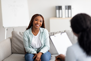 Woman is sitting on a couch, smiling, while talking to a therapist. Reaching out and talking to someone about your issues can help destress and declutter your thoughts. Reach out to a therapist at our Woodland Hills therapy office for anxiety treatment.