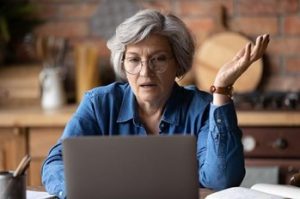 Older woman with short white hair, wearing glasses, looking at her open laptop screen in distress with her hand out.