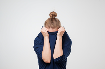 In this image we see a woman in a blue sweater standing in front of a white background. She is covering her face with her shirt. If you are afraid of failure, we offer individual therapy at our Woodland Hills therapy office that can help you overcome your fear of failing. 