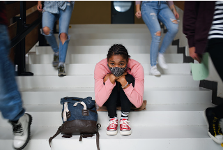 Young black girl is seen sitting on the stairs of her school, with her backpack beside her feet on the steps. There are others walking down the stairs past her as she leans her head on her knees. Teenage anxiety can get worse due to peer pressure and school stress. Adolescent therapy is offered at our Woodland Hills, CA therapy office virtually or in-person.