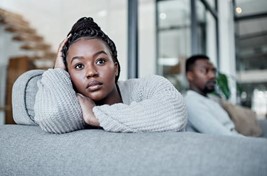 In this image we see a young black man and black woman sitting together on the couch. In the distance we see the man, and up close we see the woman turned away from the man with her hand on her head.