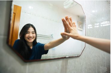 A woman is standing in front of a mirror in her bathroom, smiling at her reflection, and putting her hand on the mirror. Reframing your mindset and thoughts can help you be more positive and cope with your emotions in a healthier way. Learning coping skills and how to reframe your thoughts is something that can be worked on in individual therapy sessions with our therapists at our Woodland Hills therapy office location.