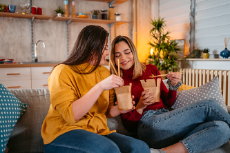 In this image we see a couple sitting on the couch, having take-out dinner together. The two woman are sitting side by side, smiling, while holding chopsticks and their food. Couples therapy is available at our therapy practice in Los Angeles, CA.