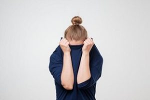In this image we see a woman in a blue sweater standing in front of a white background. She is covering her face with her shirt.