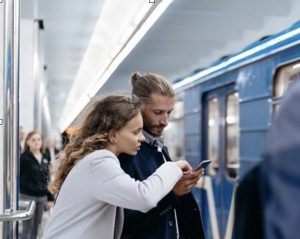 A woman leaning over a man and pointing to his phone while they stand waiting for the subway.