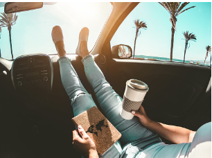 Woman in her car holding a cup of coffee, has her feet on the dashboard of her car. 