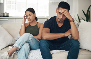 An asian-american couple sitting on the couch turning away from each other where they feel frustrated and have their hand resting on their forehead. Couple's conflict can be overwhelming. If you are feeling stuck in your relationship and learn better tools to communication, call today for our marriage counselors near you in Woodland Hills 