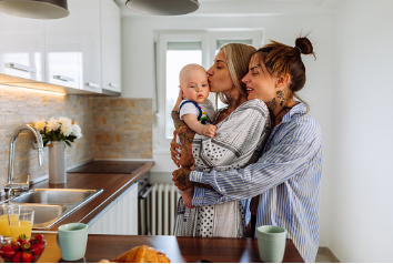 a lesbian couple standing in the kitchen and holding their baby. Parenting can create extra conflict, stress, and disconnection in a marriage. A couple's therapy can help to repair your couple's relationship so you can enjoy your family together with more joy. 91307 