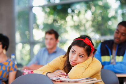 A young woman wearing a yellow shirt with a red headband sitting in a college classroom listening to a lecture and feeling stressed as she rest on her left arm. College anxiety and stress due to overwhelming responsibilities can worsen your mental health. Call today for online therapy in Chatsworth, CA. 