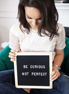 Woman holding a sign that says "Be curious, not perfect." Dr. Menije offers therapy for perfectionism at Embracing You Therapy.