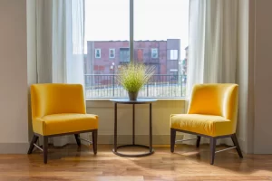 Two yellow seats in a brightly lit room with a table and plant in between them. This may represent a therapy setting.