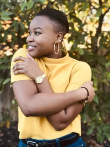 A young black woman is dressed in a yellow shirt and blue jeans, with her arms resting on her shoulders.