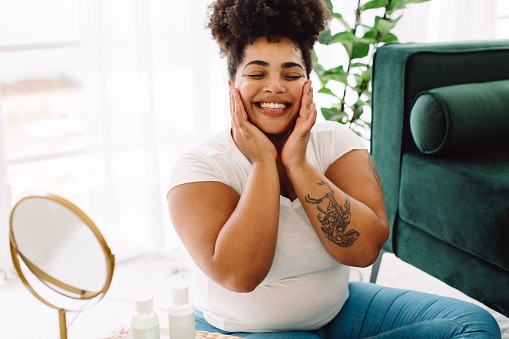 Plus size woman in living room looking happy while applying facial cream on her face. Happy female massaging her face and doing beauty care at home.