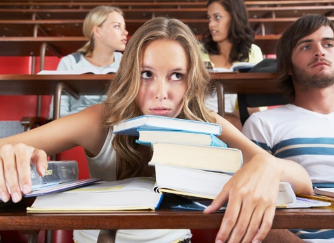A young college student sitting in a college class with pile of books that describe feeling overwhelmed and exhausted. If you are struggling with college anxiety and stress as a young adult, CBT therapists can help to teach you stress management skills. 91301 | 91302 | 91372