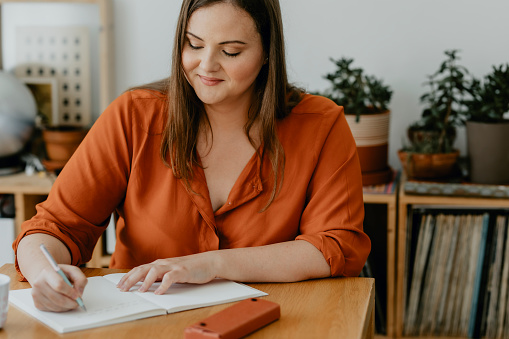 A smiling plus size woman sitting at her desk and writing a journal.