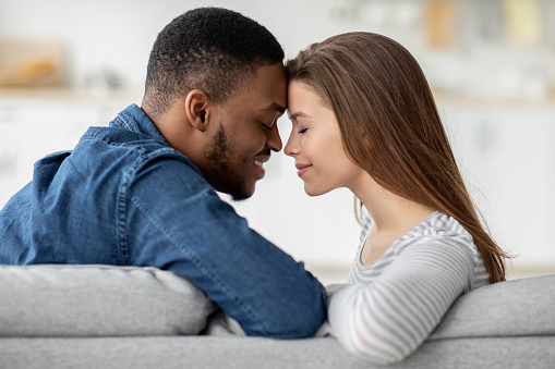 Romantic Moment. Loving Interracial Couple Bonding Together At Home, Affectionate Young Multicultural Lovers Holding Hands And Touching Foreheads While Sitting On Couch In Living Room, Closeup Shot