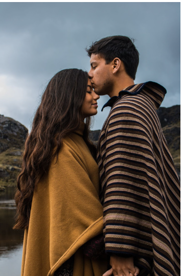 Couple is holding hands and kissing near a lake, while wearing ponchos. 