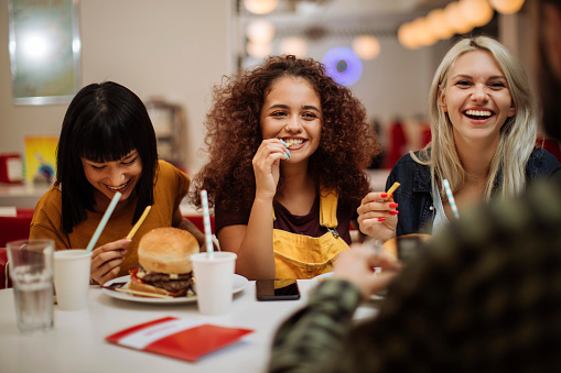 Three beautiful female friends laughing while eating burgers and fries in a restaurant. You can get help for depression and anxiety by learning CBT tools like thought-recording from a counselor in Woodland Hills, CA here in person or online therapy in California. 91335 | 91406 | 91405 | 91343
