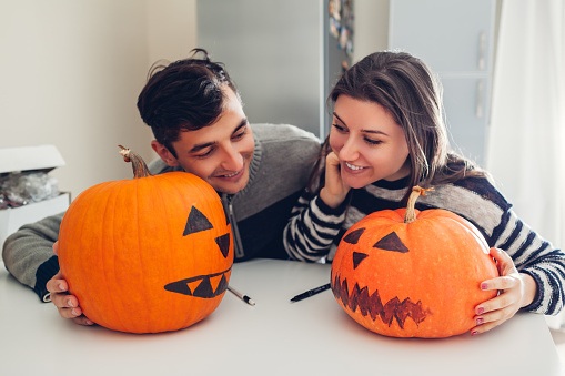 Young couple making jack-o-lantern for halloween on kitchen. Happy man and woman comparing their pumpkins