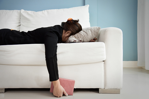 Exhausted, Tired Asian Business woman in black shirt holding notebook and sleeping on white sofa with blue wall. Stress from overtime working concept.
