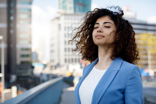 Businesswoman with curly brown hair standing outdoors in the city. The woman has her eyes closed and is wearing a white shirt with a light blue blazer. She is enjoying the breeze of the city as it blows across her.