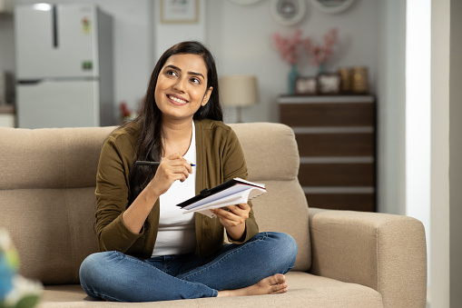 Indian American woman is sitting on her beige couch at home with her legs crossed. She is wearing blue jeans with a white shirt and a brown cardigan. She has long brown hair and is smiling while holding a pen in one hand and a notebook in the other hand.