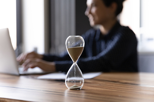 Close up hourglass measuring time, standing on wooden office table, a  businesswoman working on background, efficiency, deadline and time management concept, busy employee using laptop