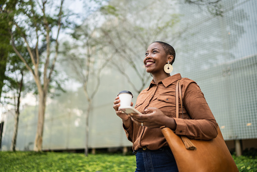 An African American woman is standing outside, wearing a brown long sleeve collared shirt. The woman is wearing large earrings and has her brown purse hanging from her shoulder. She is holding a cup of coffee in one hand and her phone in the other hand. The woman is looking up and smiling. Behind her are green trees.