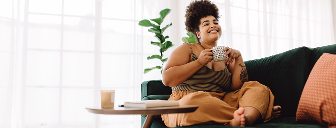 Happy female holding coffee mug while sitting on couch at home. Smiling woman sitting on sofa having coffee. If you want to improve your self-esteem, learn to forgive yourself and feel good about yourself, CBT therapy can help in Woodland Hills, CA, call today for online sessions. 91364