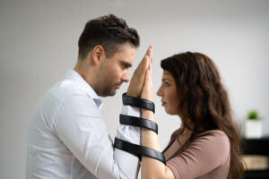 In the photo we see a man and woman facing each other. The couple both have one of their hands up on the other persons hand. Their hands are tied together with black straps, representing their codependent relationship.