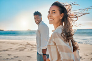 A man and woman are seen walking together on a beach, shoreside. The woman is closer to the frame, smiling as the wind blows through her brown long hair. Her partner is standing further than her, holding her hand. He is looking at her and smiling.