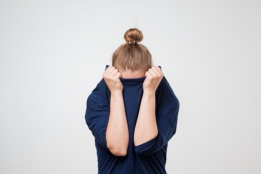 A woman is seen wearing a blue sweater, pulling the sweater over her face with both of her hands. She has blonde hair, and her hair is tied in a bun. She is displaying behaviors of wanting to escape and be alone.