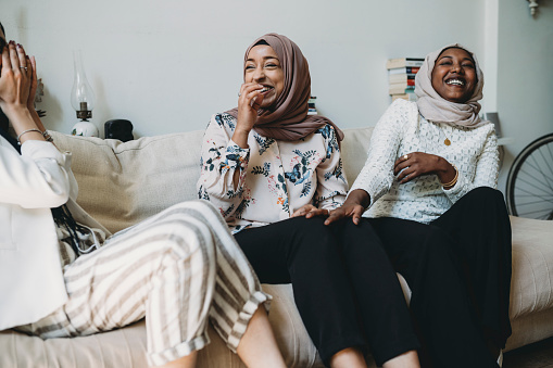 In this image we see three female friends sitting together on a couch. They are all laughing and smiling with one another. Two of the woman in the photo are wearing hijabs on their heads.