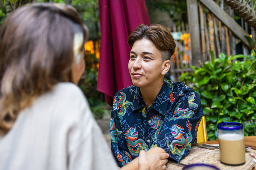 A same-sex couple is on a date at a beautiful outdoors restaurant. They are both looking at one another, admiring each other. One of the partners is backwards towards the camera, but is embracing the other woman. The woman facing the camera is smiling back at her.