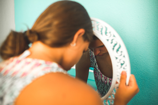 A young girl is holding a mirror and is resting her forehead on it. She is looking down and has a sad facial expression.