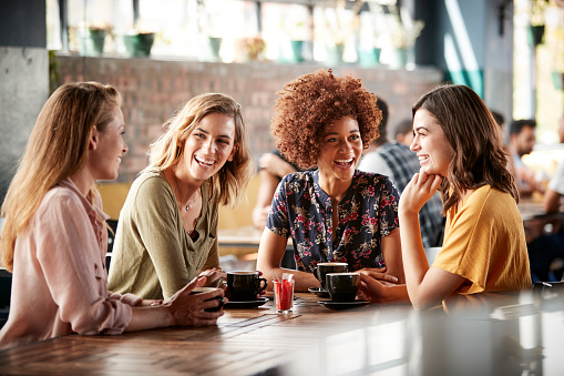 There are four young woman seated on a table at a coffee shop. They each have coffee mugs in front of them, as they smile at one another. 