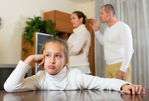 In this image we see a woman and man, with their young daughter in a living room. The young girl is seated on the floor, with her hand holding up her face. She has a sad facial expression. Her parents are behind her arguing, and not paying attention to her. All three of them are wearing white long sleeve shirts.